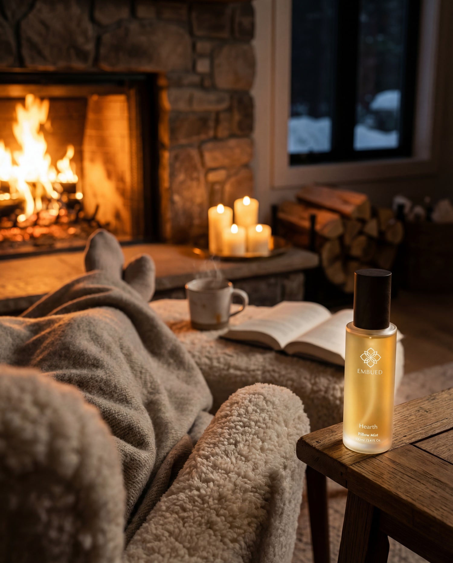 Hearth pillow mist on a timber side table beside a lit stone fireplace, with a knit blanket, candles and snow outside the window