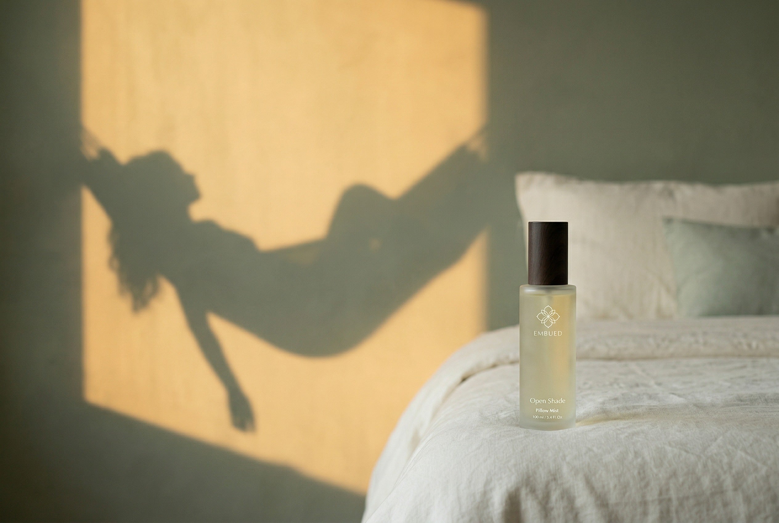 Open Shade pillow mist on a linen bed, with the shadow of a figure resting in a hammock cast across the bedroom wall