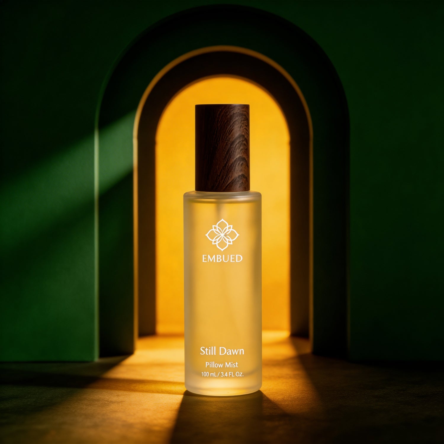 Still Dawn pillow mist by Embued — frosted 100ml glass bottle with a dark wood cap, lit by warm light in a green arched alcove