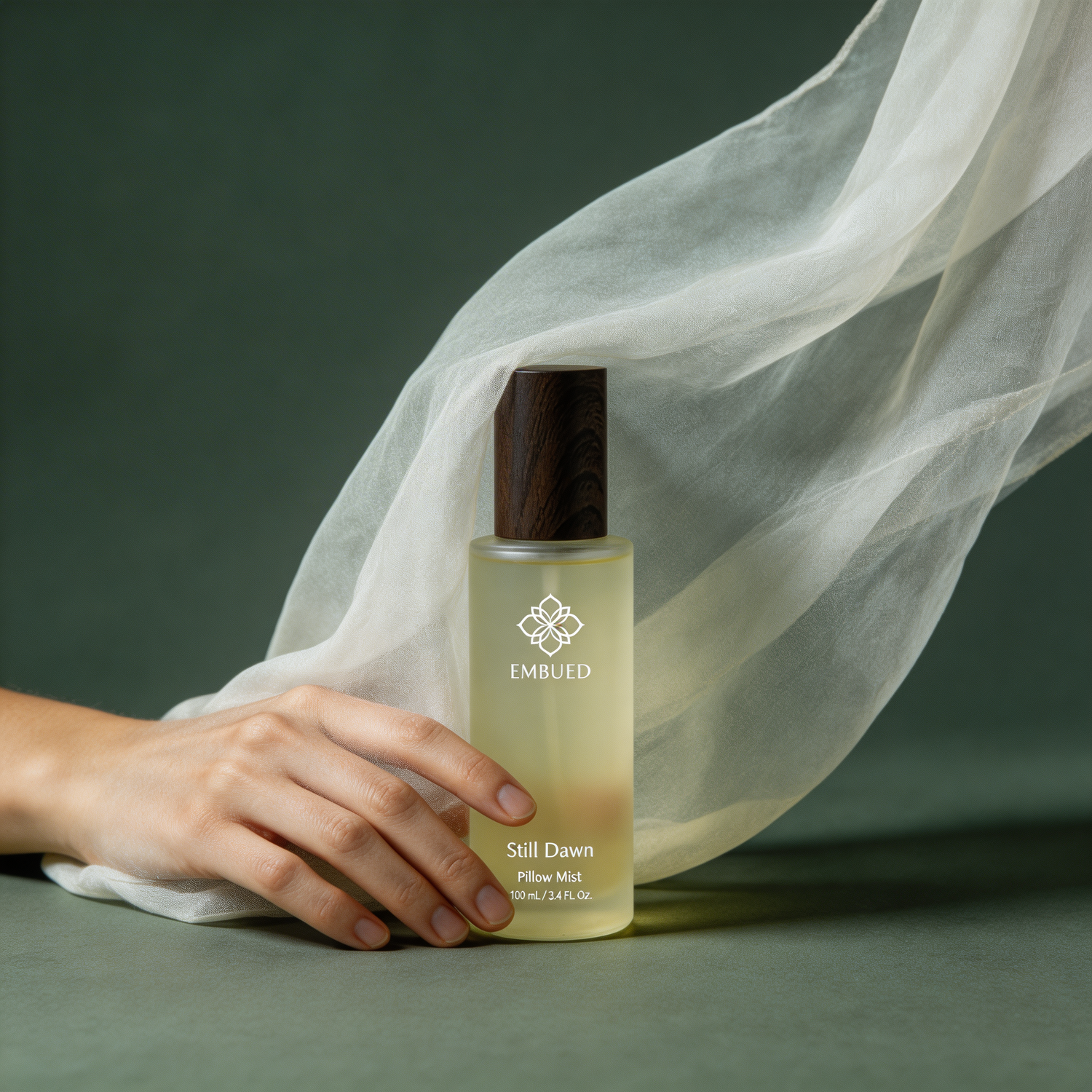 A hand resting beside the Still Dawn pillow mist, sheer fabric drifting behind the frosted 100ml bottle against deep green