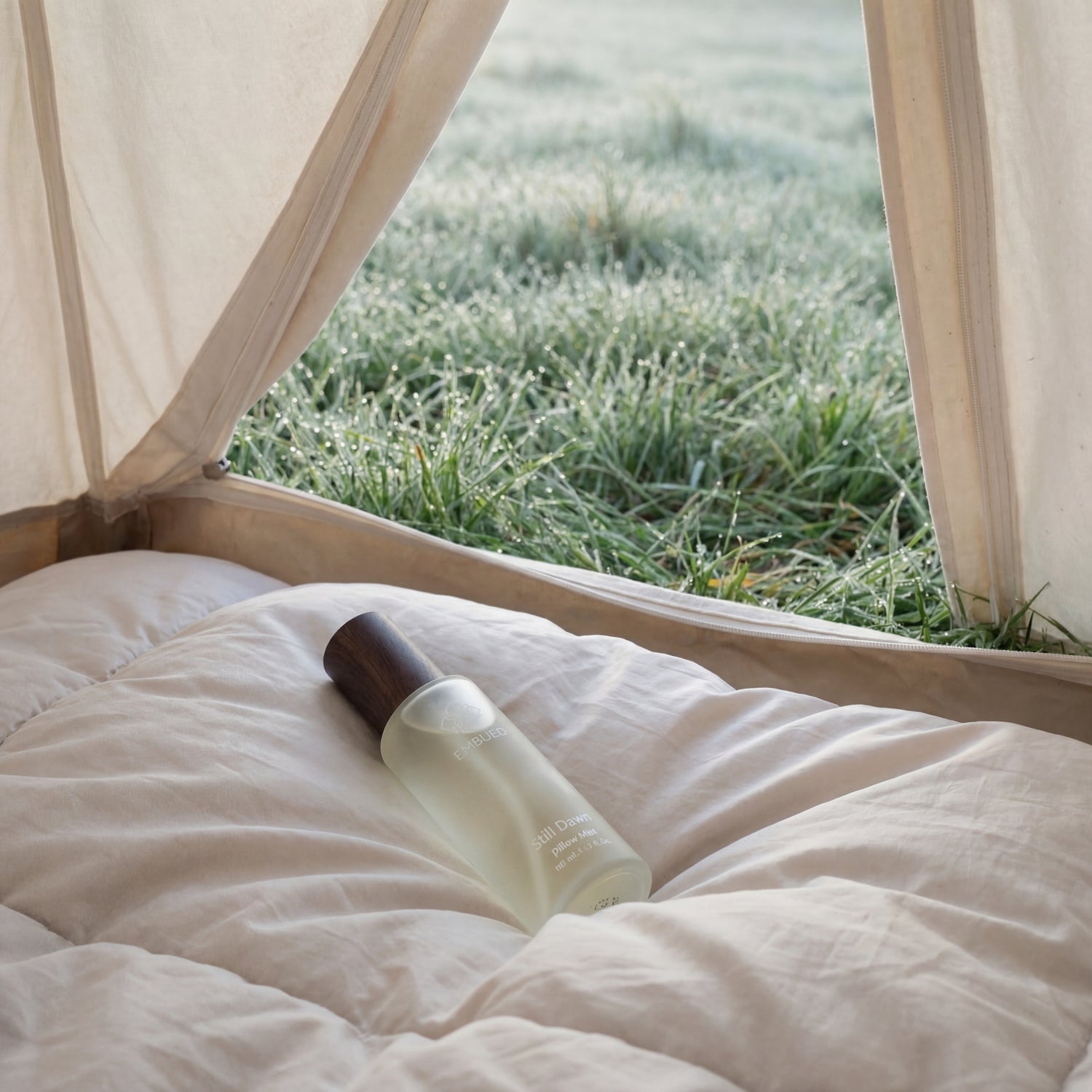 Still Dawn pillow mist resting on linen bedding inside a tent, dewy morning grass visible through the open flap